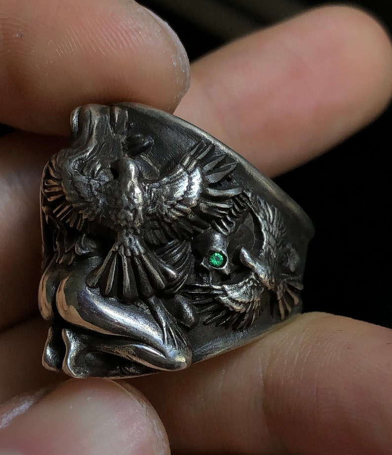 May include: A silver ring with a detailed design of a woman and two birds. The ring features a green gemstone.