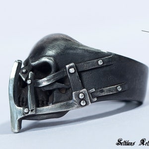 May include: A silver skull ring with a detailed design of a skull with a cage over the face. The ring is made of metal and has a black finish.