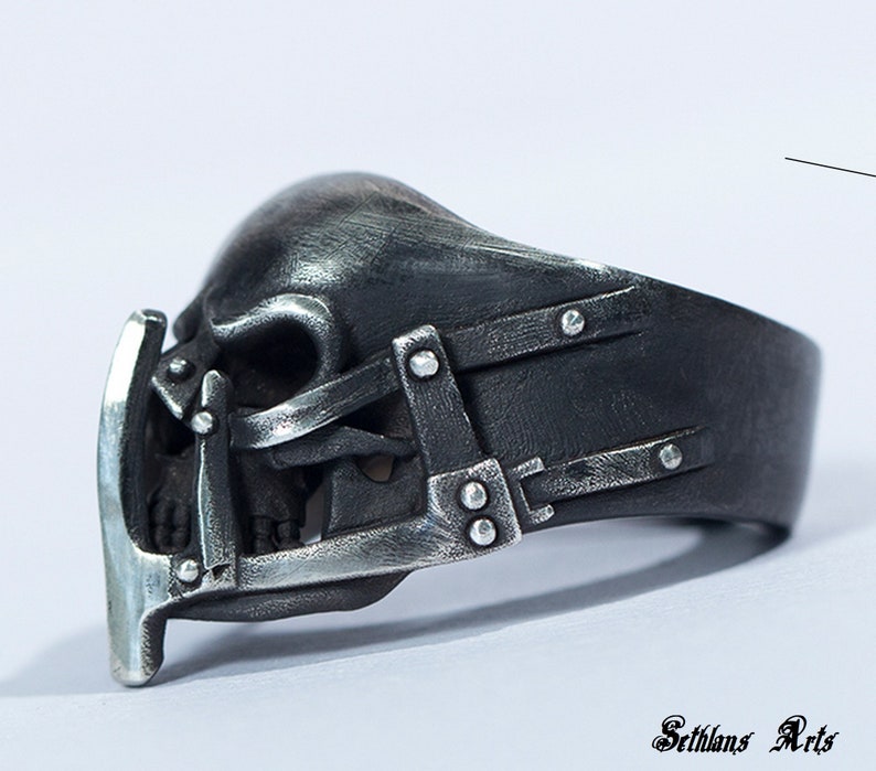 May include: A silver skull ring with a detailed design of a skull with a cage over the face. The ring is made of metal and has a black finish.