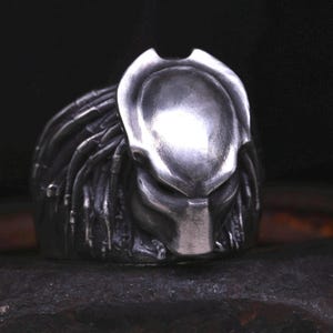 May include: A silver ring with a detailed Predator head design. The ring features a textured, sculpted head with dreadlocks.