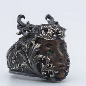 May include: A silver ring with a detailed design featuring a face with green eyes. The face is surrounded by intricate floral patterns.