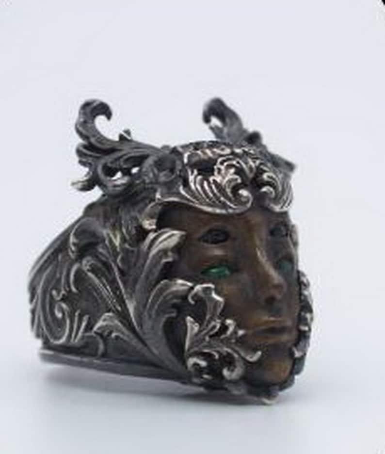 May include: A silver ring with a detailed design featuring a face with green eyes. The face is surrounded by intricate floral patterns.