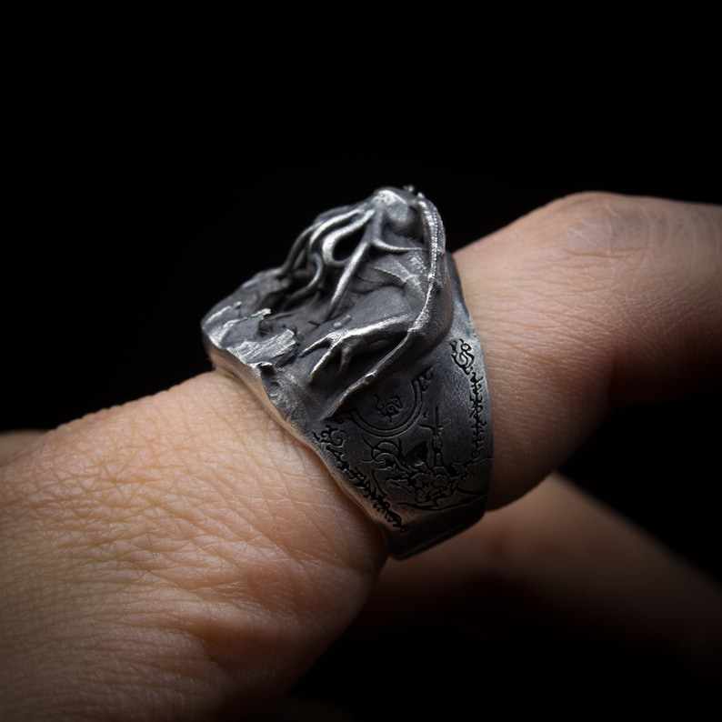 May include: A silver ring with a detailed design of a creature with tentacles. The ring is worn on a finger and has an inscription around the band.