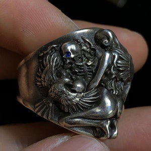 May include: A silver ring with a detailed design of a woman, a skull, and a bird. The skull has a blue gemstone in its eye socket.