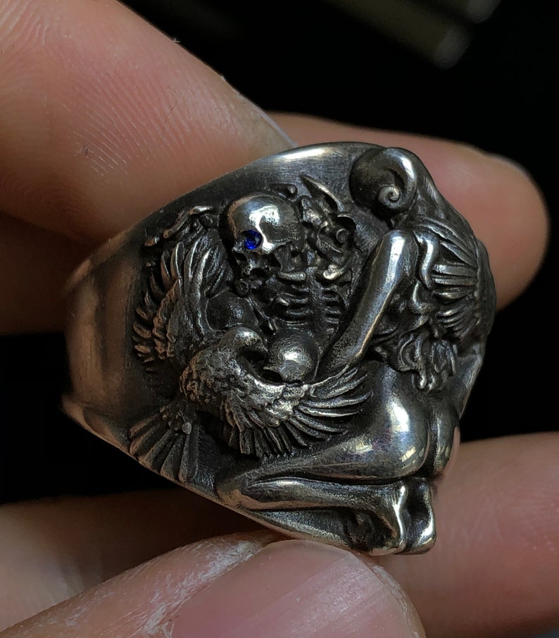 May include: A silver ring with a detailed design of a woman, a skull, and a bird. The skull has a blue gemstone in its eye socket.