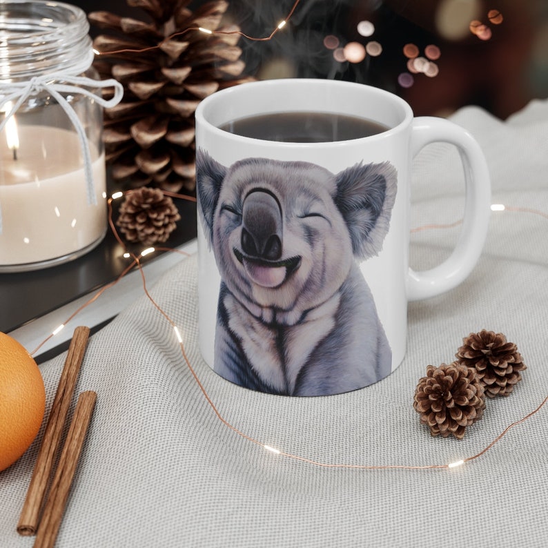 May include: White ceramic mug with a smiling koala illustration. The koala has a light gray fur and is shown with its tongue sticking out. The mug is filled with coffee.