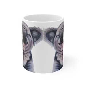 May include: White ceramic mug with a smiling koala face printed on both sides. The koala has a tongue sticking out and its eyes are closed.