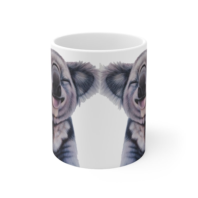 May include: White ceramic mug with a smiling koala face printed on both sides. The koala has a tongue sticking out and its eyes are closed.