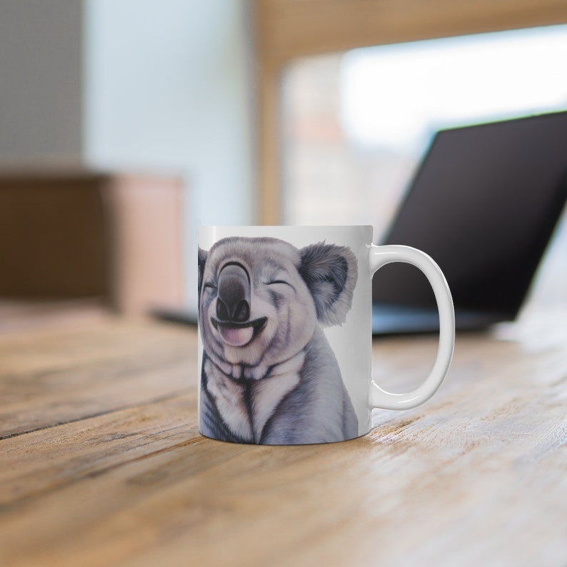May include: White ceramic mug with a smiling koala printed on it. The koala is gray and has a black nose and eyes.
