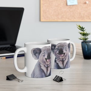 May include: Two white ceramic mugs with a smiling koala illustration on each. The koalas are drawn in a realistic style with gray fur and black eyes. The mugs are a standard size and have a handle on the right side.