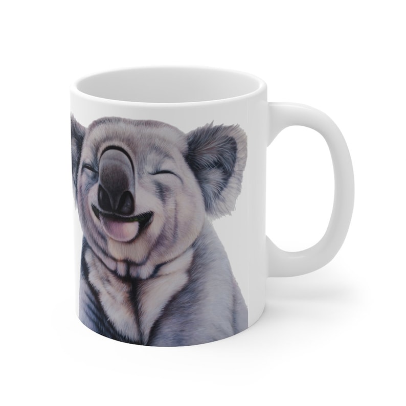 May include: White ceramic coffee mug with a smiling koala illustration. The koala has closed eyes and a big grin, with its tongue sticking out.