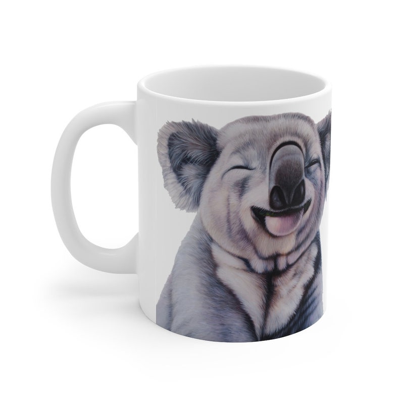 May include: White ceramic mug with a smiling koala illustration. The koala has closed eyes and its tongue is sticking out. The koala is drawn in a realistic style with shades of gray and white.