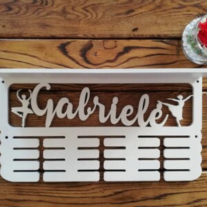 May include: White wooden medal display with the name "Gabriele" and two ballerina silhouettes. The display has 16 slots for medals.