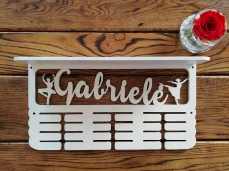 May include: White wooden medal display with the name "Gabriele" and two ballerina silhouettes. The display has 16 slots for medals.