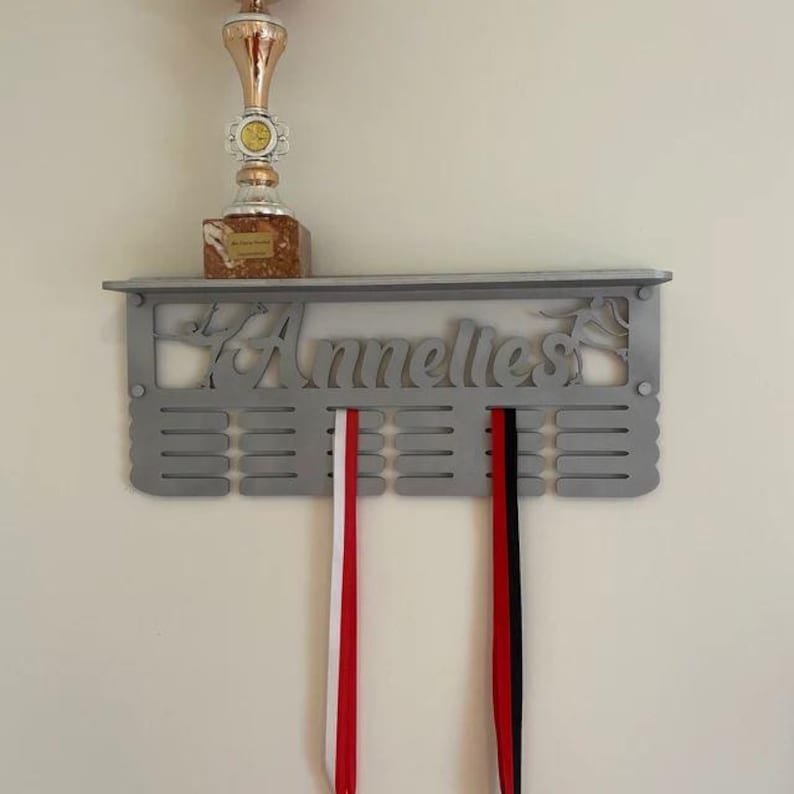 May include: A silver medal display rack with the name "Annelies" engraved on it. The rack has 12 slots for medals and a shelf on top. Two red and white striped ribbons are hanging from the slots.