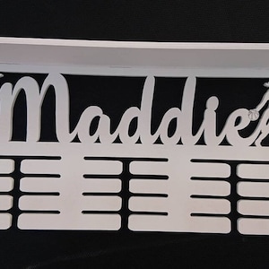 May include: White wooden medal display with the name Maddie cut out. The display has two figures, one playing baseball and the other playing basketball. There are 12 slots for medals.