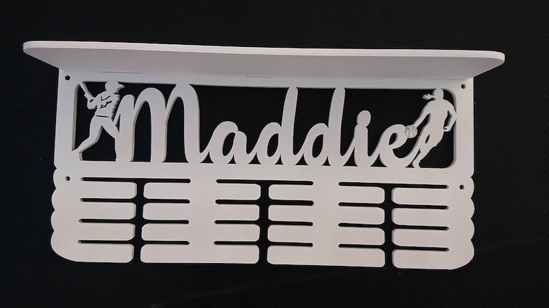 May include: White wooden medal display with the name Maddie cut out. The display has two figures, one playing baseball and the other playing basketball. There are 12 slots for medals.