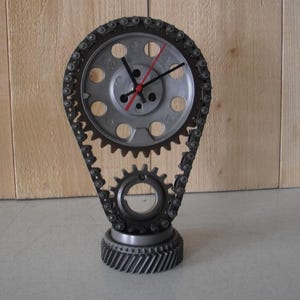 May include: A unique clock made from repurposed metal gears and a timing chain. The clock face is silver with black numbers and hands, and a red second hand. The clock stands on a gear base, creating an industrial, mechanical aesthetic.