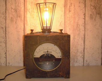 Könnte beinhalten: Eine Tischlampe im Vintage-Stil aus einer wiederverwendeten Holzkiste mit einem runden analogen Messgerät mit der Aufschrift "AMPERE" und einer Käfiglampe. Die Lampe hat ein schwarzes Kabel und einen Stecker.