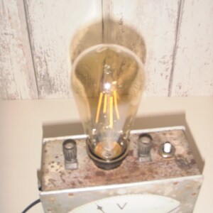 K&ouml;nnte beinhalten: Eine Lampe im Vintage-Stil, hergestellt aus einem wiederverwendeten Voltmeter. Die Lampe hat eine klare Gl&uuml;hbirne mit einem Filament und ein wei&szlig;es Zifferblatt mit einem schwarzen Zeiger und dem Buchstaben "V" f&uuml;r Volt. Die Lampe steht auf einer Holzoberfl&auml;che.