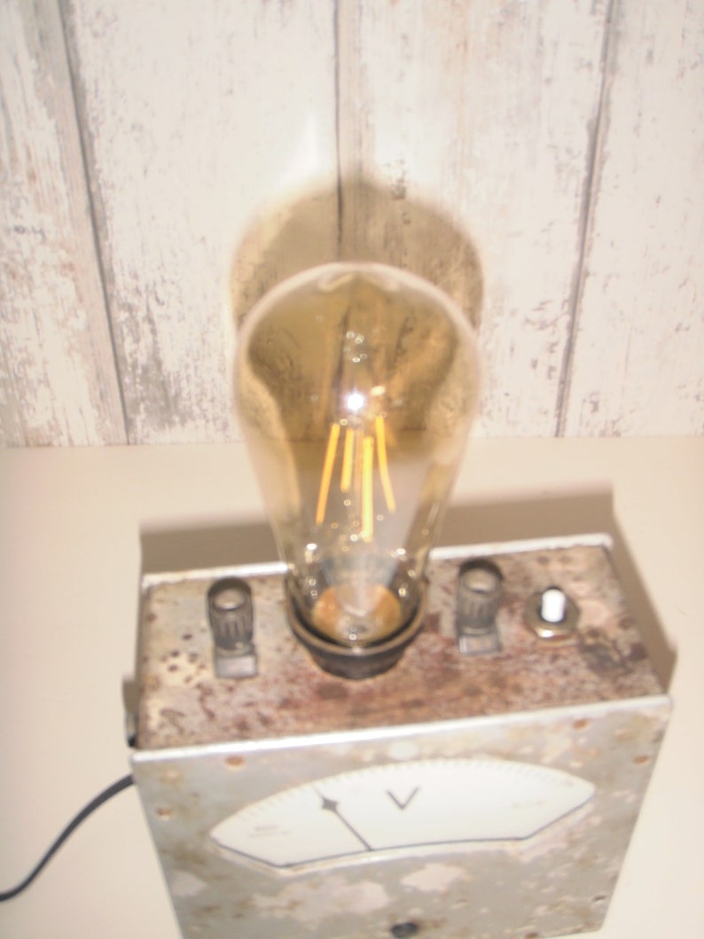 K&ouml;nnte beinhalten: Eine Lampe im Vintage-Stil, hergestellt aus einem wiederverwendeten Voltmeter. Die Lampe hat eine klare Gl&uuml;hbirne mit einem Filament und ein wei&szlig;es Zifferblatt mit einem schwarzen Zeiger und dem Buchstaben "V" f&uuml;r Volt. Die Lampe steht auf einer Holzoberfl&auml;che.