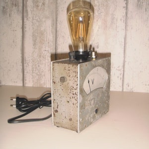K&ouml;nnte beinhalten: Ein Vintage-Elektroger&auml;t aus grauem Metall mit einem runden Analogmessger&auml;t und einer Gl&uuml;hbirne. Das Ger&auml;t hat ein schwarzes Kabel und einen Stecker. Das Messger&auml;t hat eine Nadel, die auf den Buchstaben "V" und die Zahl "2" zeigt.