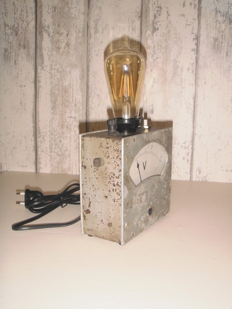 K&ouml;nnte beinhalten: Ein Vintage-Elektroger&auml;t aus grauem Metall mit einem runden Analogmessger&auml;t und einer Gl&uuml;hbirne. Das Ger&auml;t hat ein schwarzes Kabel und einen Stecker. Das Messger&auml;t hat eine Nadel, die auf den Buchstaben "V" und die Zahl "2" zeigt.