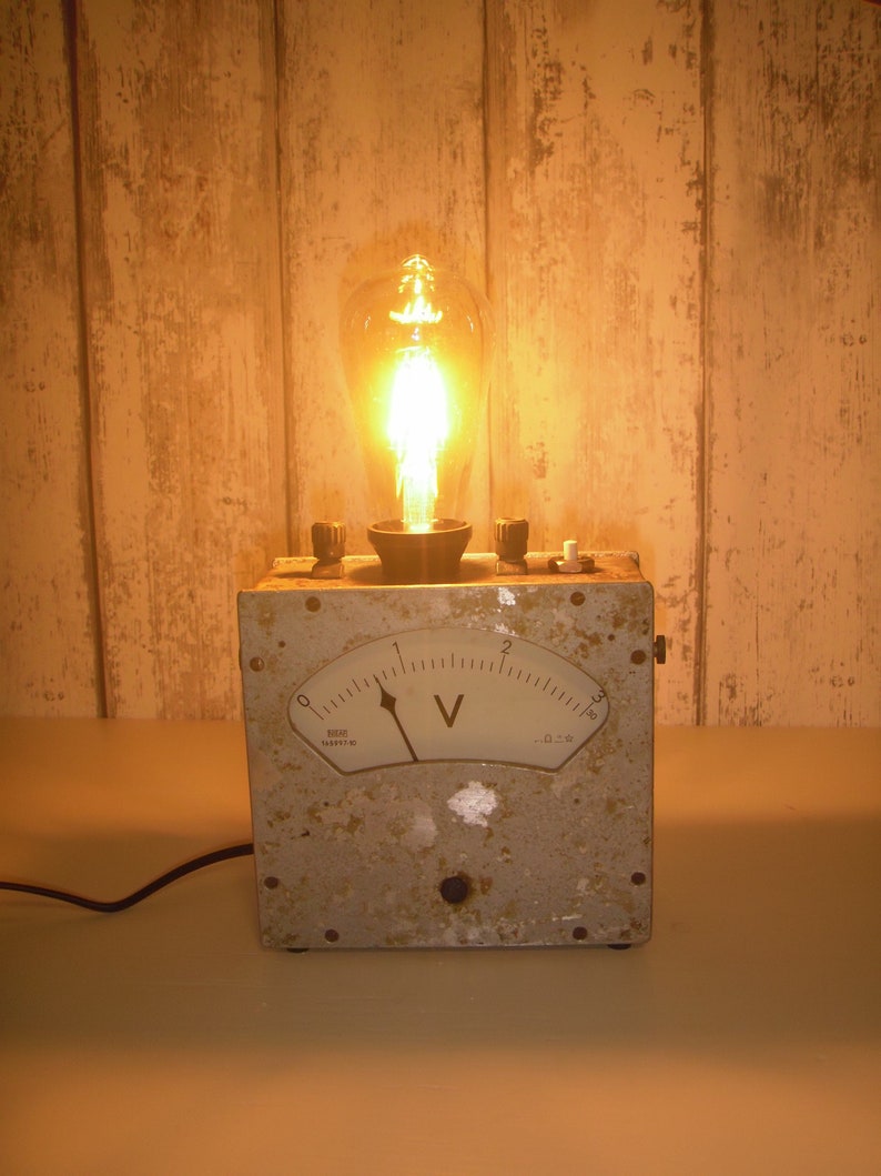 K&ouml;nnte beinhalten: Eine Lampe im Vintage-Stil, hergestellt aus einem wiederverwendeten Voltmeter. Die Lampe hat eine klare Gl&uuml;hbirne mit einem gl&uuml;henden Filament und ein wei&szlig;es Zifferblatt mit schwarzen Markierungen. Das Voltmeter zeigt "V" an und hat Zahlen von 0 bis 3. Die Lampe steht auf einer hellbraunen Holzoberfl&auml;che.