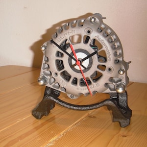May include: A unique clock made from a repurposed automotive alternator. The clock features a silver-colored metal casing with visible gears and bolts, a black clock face, and red and black hands. The clock is mounted on a black metal stand.