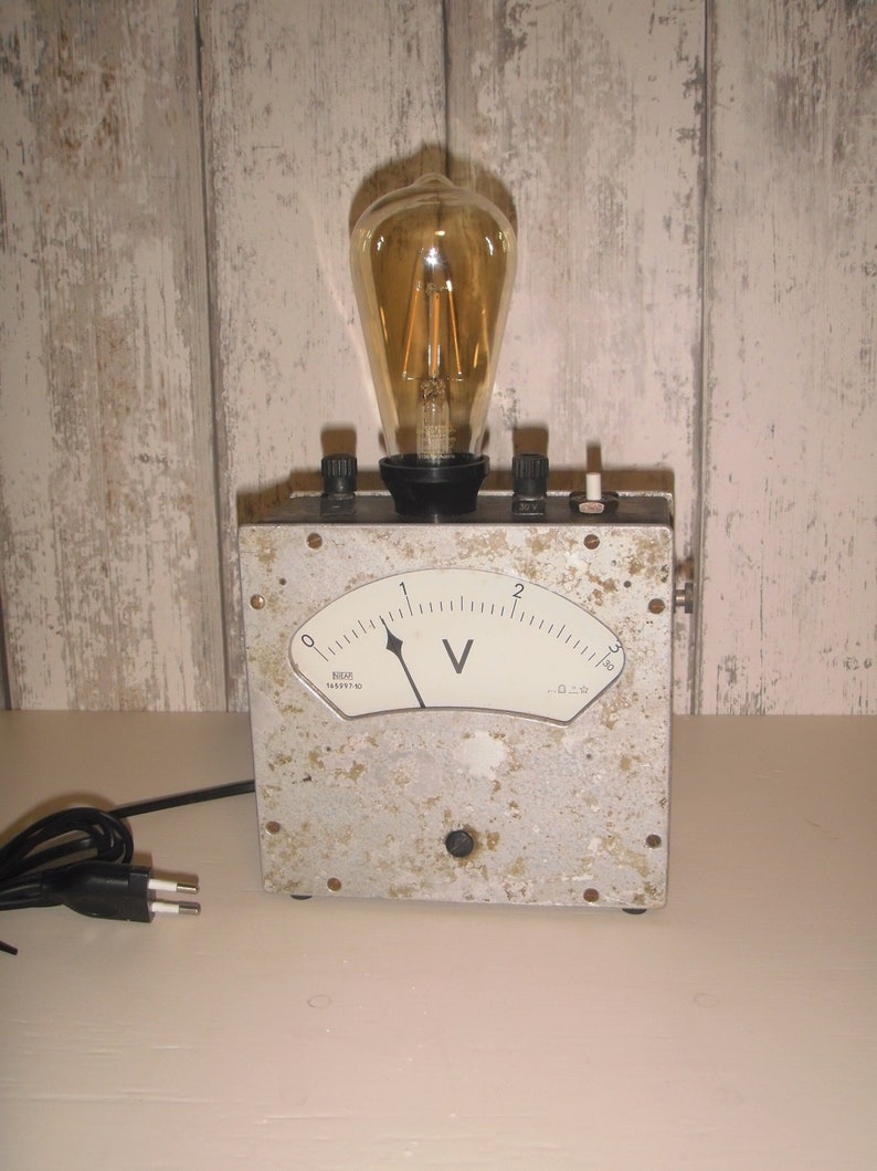 K&ouml;nnte beinhalten: Ein analoges Voltmeter im Vintage-Stil mit einer klaren Gl&uuml;hbirne oben. Das Voltmeter hat ein wei&szlig;es Zifferblatt mit schwarzen Markierungen und einem schwarzen Zeiger. Die Gl&uuml;hbirne leuchtet und hat einen warmen Schein. Das Ger&auml;t ist aus Metall und hat eine graue Oberfl&auml;che. Es hat ein schwarzes Stromkabel mit einem Stecker.