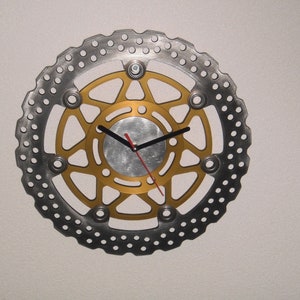 May include: A gold and silver clock with a circular design. The clock face is made of a metal disc with a starburst pattern. The clock hands are black and red.