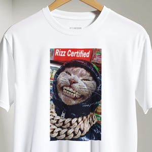 May include: White t-shirt with a smiling cat wearing a black hoodie and a gold chain. The cat image is set against a red background with the text "Rizz Certified". The shirt is hanging on a wooden hanger.