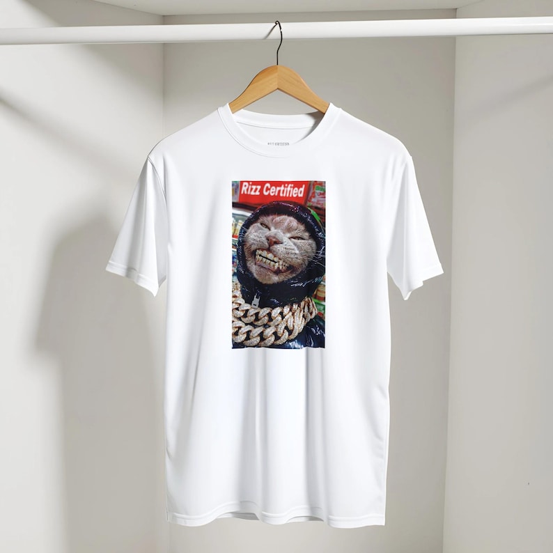May include: White t-shirt with a smiling cat wearing a black hoodie and a gold chain. The cat image is set against a red background with the text "Rizz Certified". The shirt is hanging on a wooden hanger.
