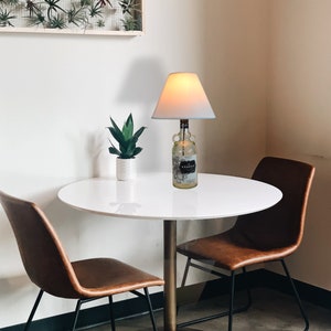 Puede incluir: Una lámpara de mesa hecha de una botella reutilizada, con una pantalla blanca y un borde azul. La lámpara está sobre una mesa redonda blanca, con dos sillas de cuero marrón. También se ve una pequeña planta en maceta y una decoración de pared enmarcada.