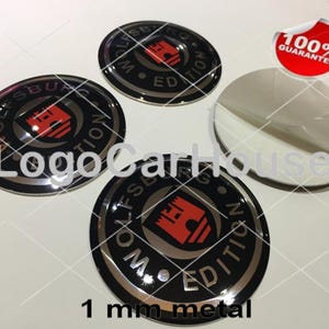 May include: Three circular metal emblems featuring a black background with a silver border and the text Wolfsburg Edition surrounding a red castle logo. One emblem is shown with its adhesive backing partially peeled off. Includes a 100% guarantee sticker.