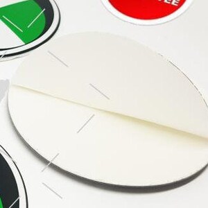 May include: A close-up of a white, folded, oval-shaped sticker. The sticker is partially peeled back, revealing a crease. Other stickers with green, black, and red accents are visible in the background.