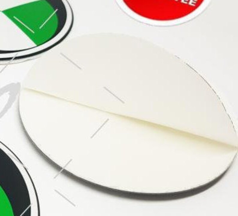 May include: A close-up of a white, folded, oval-shaped sticker. The sticker is partially peeled back, revealing a crease. Other stickers with green, black, and red accents are visible in the background.