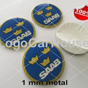 Könnte beinhalten: Mehrere runde Metall-Embleme in Blau und Gold mit dem Wort "SAAB" und drei goldenen Kronen. Die Embleme sind 1 mm dick. Ein rot-weißer Aufkleber mit dem Text "100% GARANTIE" ist ebenfalls sichtbar.