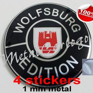 Op de afbeelding: Een rond zwart en zilverkleurig metalen embleem met de tekst WOLFSBURG EDITION en een rood kasteelpictogram in het midden. Een kleine rode ronde badge in de rechterbovenhoek vermeldt 100% GUARANTEE. Een kleine inzet toont een witte zelfklevende achterkant.