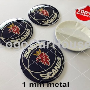 Könnte beinhalten: Vier 1 mm dicke Scania-Autoembleme aus Metall mit blauem Hintergrund, silbernem Scania-Logo und einem roten und goldenen Greif. Die Embleme befinden sich auf einer weißen Oberfläche mit einem 100% Garantie-Aufkleber.