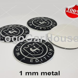 Könnte beinhalten: Drei kreisförmige schwarze und silberne Metallembleme mit dem Text Wolfsburg Edition und einer zentralen Grafik aus Burg und Wolf. Ein Emblem ist umgedreht und zeigt eine weiße Kleberückseite. Der Text 1 mm metal ist unten zu sehen.