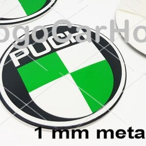 May include: Circular metal emblem with the word "PUCH" in white, surrounded by a black border. The emblem features a green and white checkered design. The text "1 mm metal" is at the bottom. The emblem is likely for a vehicle.