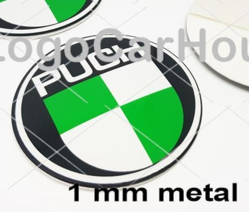 May include: Circular metal emblem with the word "PUCH" in white, surrounded by a black border. The emblem features a green and white checkered design. The text "1 mm metal" is at the bottom. The emblem is likely for a vehicle.