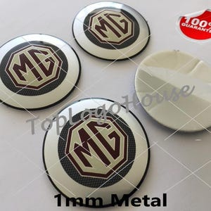 May include: Three circular MG logo emblems with a white background, a dark red and cream octagonal center, and a textured black border. One additional circular emblem is shown with its white adhesive backing facing upward. Text reads 100% GUARANTEE.