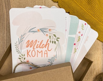 May include: A brown cardboard box containing a stack of rectangular cards with rounded corners. The top card features a floral wreath illustration and the text "Milch KOMA" in orange script. A small ball of brown twine sits next to the open box.