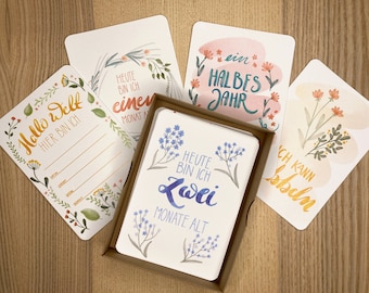 May include: A set of rectangular milestone cards with watercolour floral illustrations and German text. One card in a brown box reads "HEUTE BIN ICH ZWEI MONATE ALT" with blue flowers. Other cards feature "Hallo Welt", "ein HALBES JAHR", and "ICH KANN krabbeln".