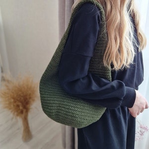 Puede incluir: Un bolso tote de crochet verde con un diseño holgado. El bolso se lleva sobre el hombro de una persona que lleva una camisa y pantalones negros.