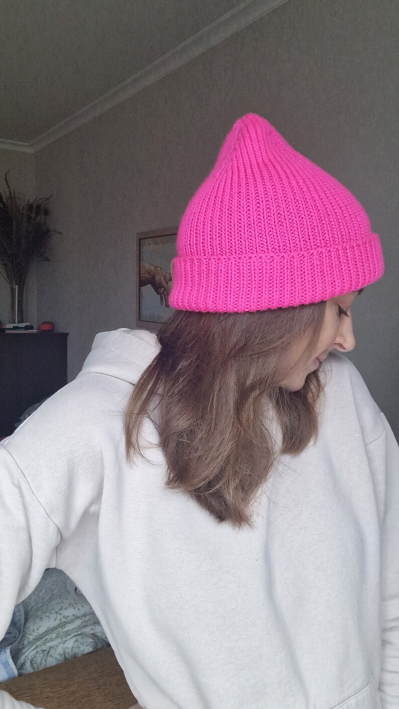 Op de afbeelding: Een felroze, geribbelde gebreide beanie. De hoed heeft een levendige fuchsia kleur en heeft een puntige bovenkant. De hoed is gemaakt van een dik, warm materiaal, perfect voor koud weer. De hoed is een stijlvol accessoire.