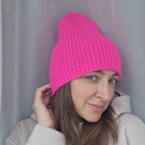 Op de afbeelding: Een felroze, geribbelde gebreide beanie. De hoed heeft een levendige, effen kleur en een klassieke, losse pasvorm. De hoed is gemaakt van een zacht, warm materiaal, ideaal voor koud weer.