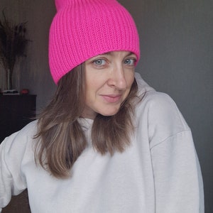 Op de afbeelding: Een felroze, geribbelde gebreide beanie. De hoed heeft een effen kleur en een losse pasvorm. De hoed wordt gedragen met een witte hoodie.